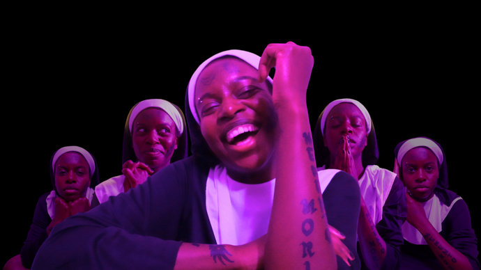 Person dressed in a nun's habit with visible tattoos on their face and arm, smiling with other similarly dressed figures in varied poses against a black background.