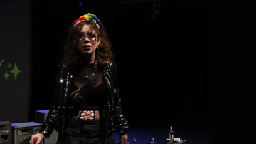 Woman with brown curly hair and technicolour hair band wears black leather jacket and looks to the camera