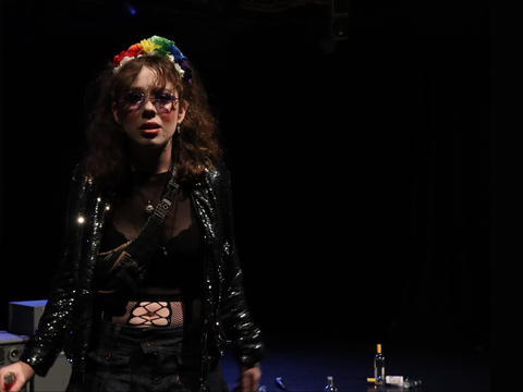 Woman with brown curly hair and technicolour hair band wears black leather jacket and looks to the camera