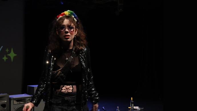 Woman with brown curly hair and technicolour hair band wears black leather jacket and looks to the camera