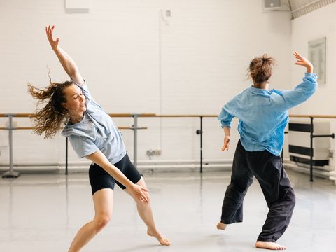 Two dancers in casual attire move gracefully in a spacious dance studio with wooden floors and white walls.