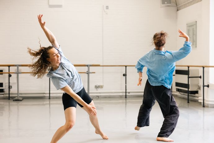 Two dancers in casual attire move gracefully in a spacious dance studio with wooden floors and white walls.