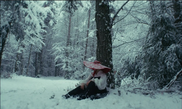 Person in a red hat and white coat sits against a tree in a snowy forest, surrounded by snow-covered trees.