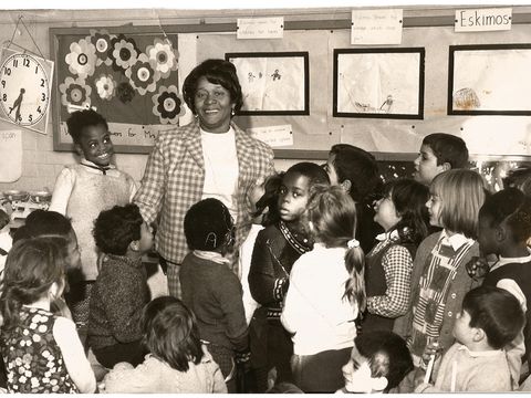 black and white photo of woman standing surrounding by children in classroom with displays behind