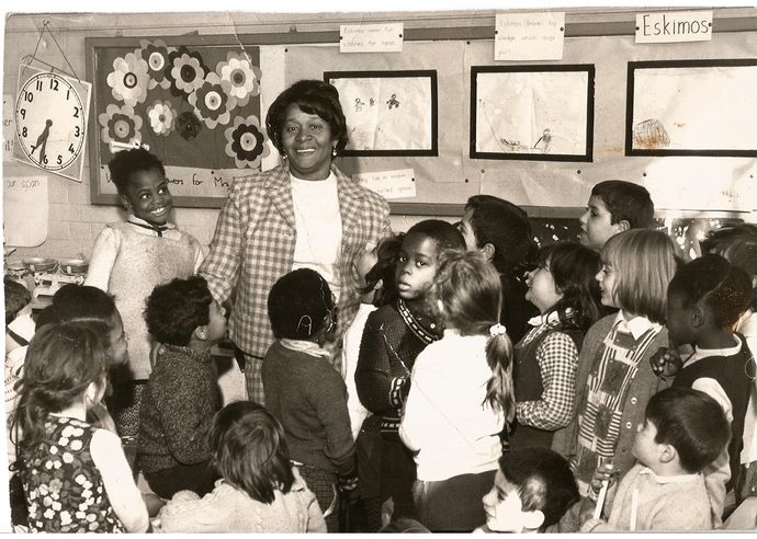 black and white photo of woman standing surrounding by children in classroom with displays behind