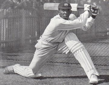A cricketer in white uniform and pads executes a batting stance, holding a cricket bat, with a netted fence in the background.