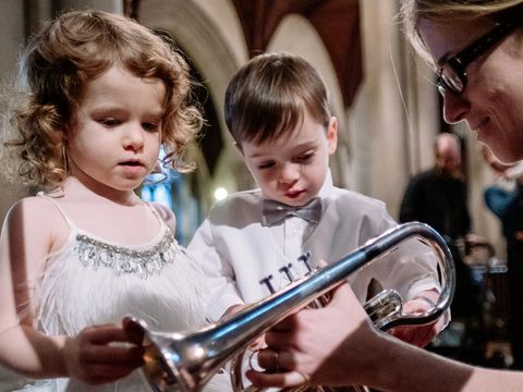 Two young children closely examine a trumpet held by an adult in a building with stone columns.
