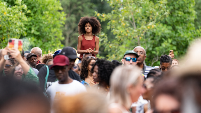 A child in a red top stands above a crowd outdoors, surrounded by trees. People are taking photos and engaging with the event.