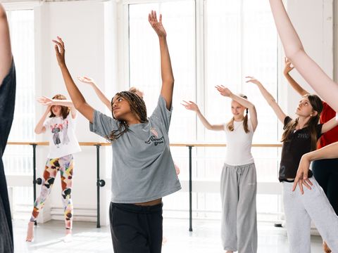 A group of people practicing dance in a bright studio, raising their arms gracefully, wearing casual clothes, and appearing focused and engaged.