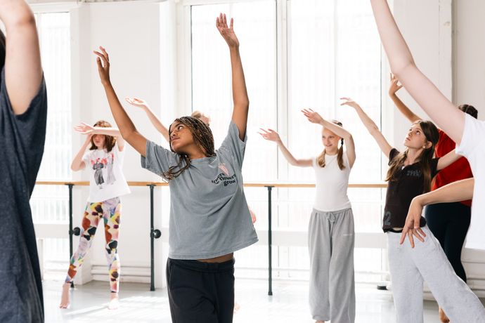 A group of people practicing dance in a bright studio, raising their arms gracefully, wearing casual clothes, and appearing focused and engaged.