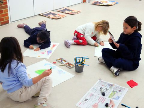 Children sitting on the floor engaged in drawing and coloring on paper with pencils and crayons, with one child using a smartphone.