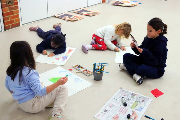 Children sitting on the floor engaged in drawing and coloring on paper with pencils and crayons, with one child using a smartphone.