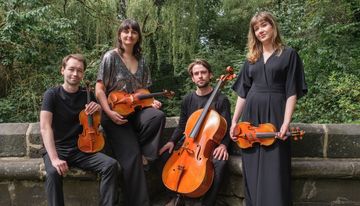 Four people stand outdoors against a stone wall, each holding a string instrument, with trees and greenery behind them.