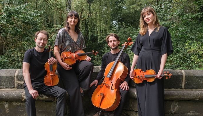 Four people stand outdoors against a stone wall, each holding a string instrument, with trees and greenery behind them.