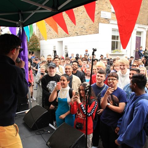 View from stage across crowd on residential street. the stage has colourful bunting hanging from the cover as a man stands on stage speaking into microphone