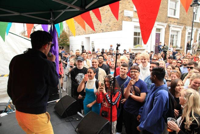 View from stage across crowd on residential street. the stage has colourful bunting hanging from the cover as a man stands on stage speaking into microphone