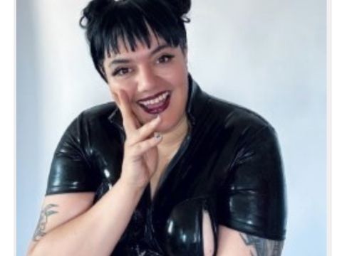 Person with dark hair in two buns and tattoos, smiling brightly, wearing a shiny black outfit, posing with a hand on their cheek.