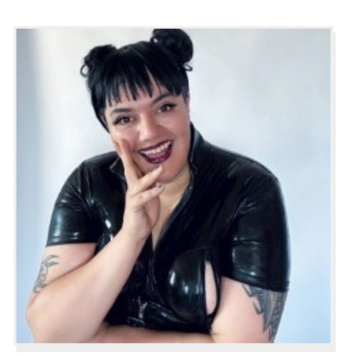 Person with dark hair in two buns and tattoos, smiling brightly, wearing a shiny black outfit, posing with a hand on their cheek.