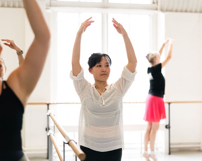 People practicing ballet in a studio, focusing on their arm positions near a barre with large windows providing natural light.