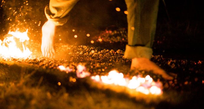 Bare feet walking over glowing embers in the dark, surrounded by small flames and sparks, creating a dramatic and intense scene.