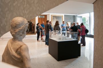 A group of people stands in a museum gallery, listening to a guide, with art pieces displayed on the walls and a sculpture in the foreground.