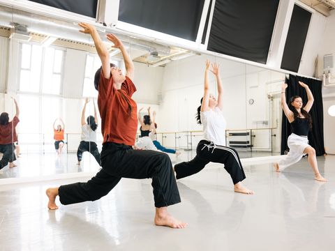 Three dancers practice a routine in a bright studio, each in a lunge position with arms raised. A mirrored wall reflects their movements.