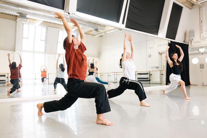 Three dancers practice a routine in a bright studio, each in a lunge position with arms raised. A mirrored wall reflects their movements.