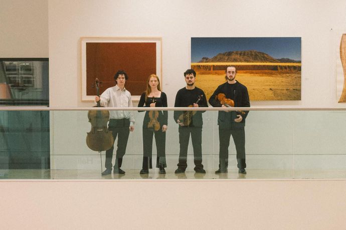Four people stand in a gallery holding string instruments, with framed art and a landscape photo on the wall behind them.