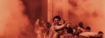 A joyous couple embraces in the foreground, surrounded by swirling orange smoke and chaotic movement in a historical setting.
