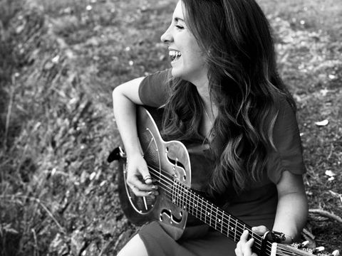 A woman smiling and playing a guitar while sitting on grass outdoors. Her long hair is down, and she appears joyful and relaxed. Black and white photo.