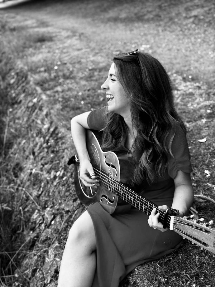 A woman smiling and playing a guitar while sitting on grass outdoors. Her long hair is down, and she appears joyful and relaxed. Black and white photo.