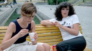 Two women sit on a yellow bench; one eats a yogurt, the other wears a Mickey Mouse T-shirt, near a waterway in a park.