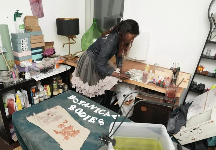 A woman works on art projects at a cluttered table in a creative studio, surrounded by various craft supplies and colorful materials.