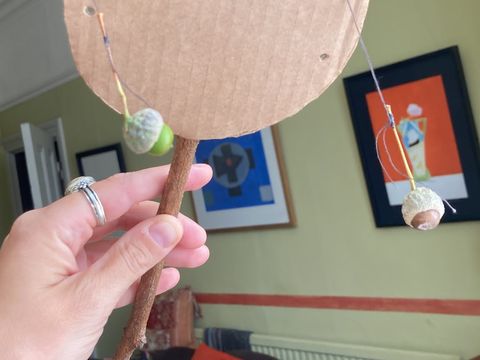 Hand holding a homemade rattle with a cardboard circle and stick handle, two small objects attached by strings. Background shows a living room with colorful art.