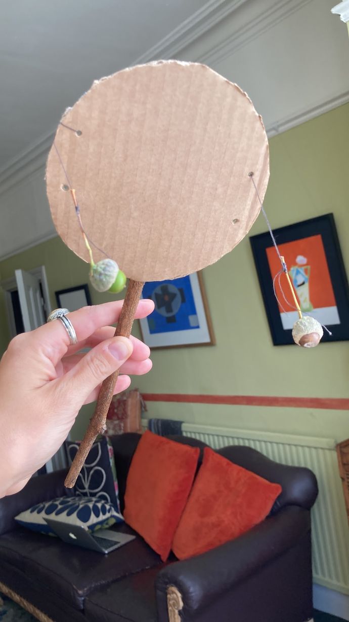 Hand holding a homemade rattle with a cardboard circle and stick handle, two small objects attached by strings. Background shows a living room with colorful art.