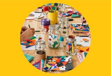 People crafting colorful paper art on a wooden table, surrounded by jars of pens and scissors, on a yellow background.