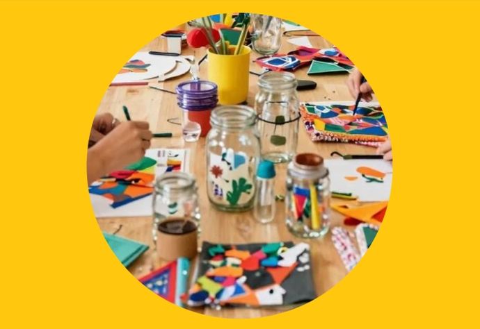 People crafting colorful paper art on a wooden table, surrounded by jars of pens and scissors, on a yellow background.