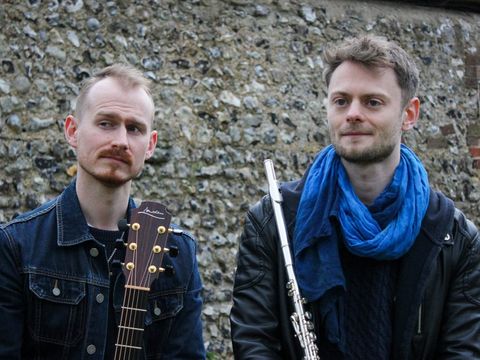 Two men stand outdoors with brick wall behind them, one is holding a guitar, the other is holding a flute or recorder
