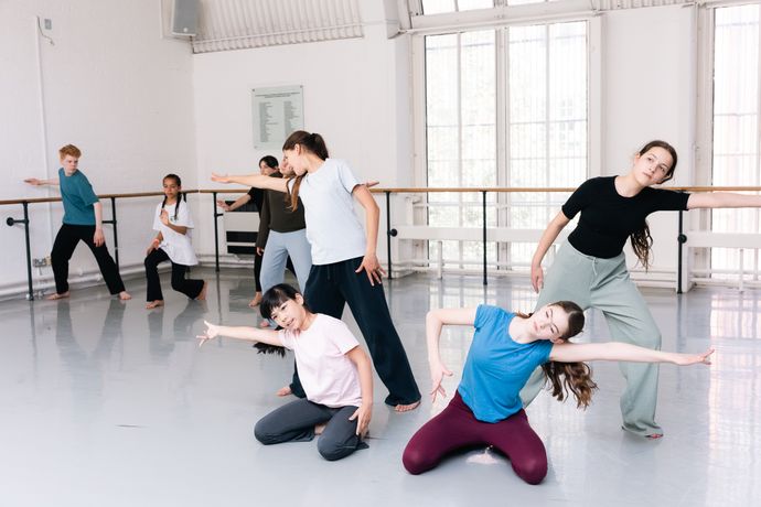 A group of students practicing contemporary dance in a studio, with some standing and others crouching, expressing dynamic movement.