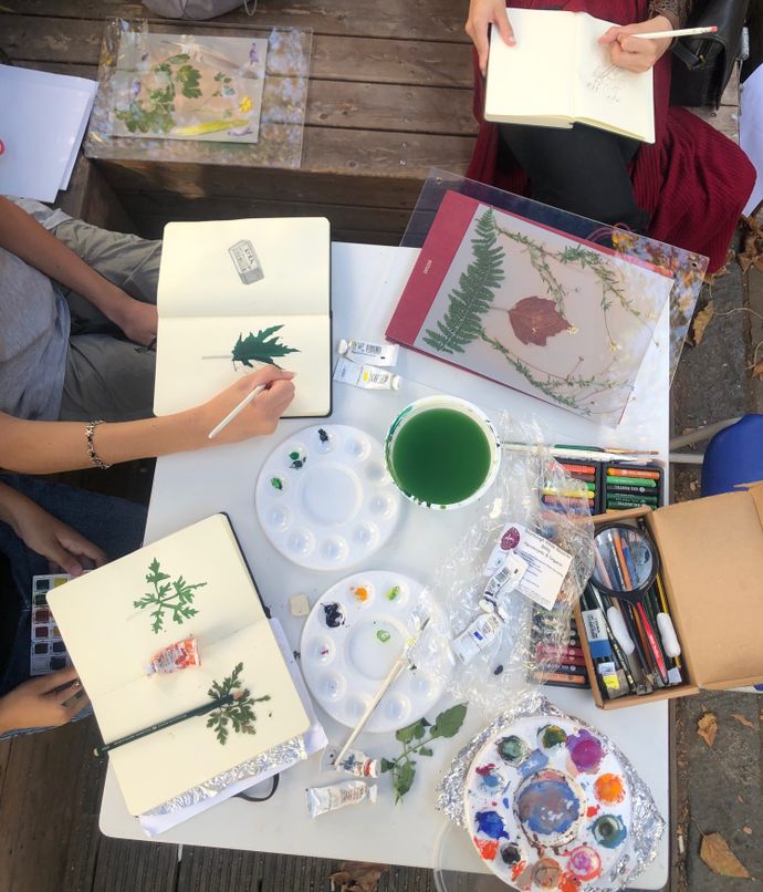 Overhead view of an outdoor painting session: open sketchbooks with leaf studies, paint palettes, brushes, green rinse cup, tubes of paint and pressed plants.