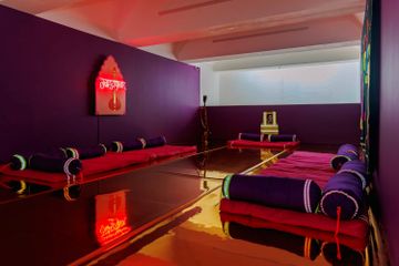 A room with purple walls, red cushions, and gold flooring. A neon sign and a framed photo are displayed, creating a serene atmosphere.