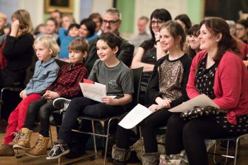 A group of children and adults sit attentively in chairs, holding papers, in a brightly lit room with an audience in the background.