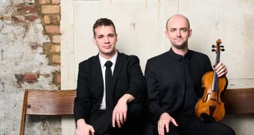 Two people dressed in formal black clothing sit on a wooden bench against a textured wall, with one person holding a violin.