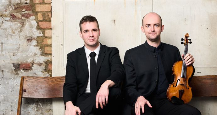 Two people dressed in formal black clothing sit on a wooden bench against a textured wall, with one person holding a violin.