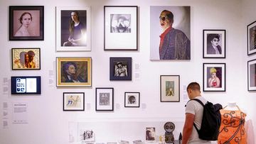 A person with a backpack observes a wall of diverse artworks, including portraits and abstract pieces, in an art gallery exhibit.