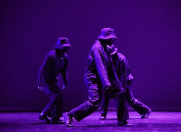 Dancers in matching outfits and hats perform on stage under purple lighting.