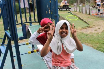 Two children smiling and making peace signs on a playground slide, with outdoor seating and play equipment in the background.