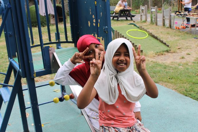Two children smiling and making peace signs on a playground slide, with outdoor seating and play equipment in the background.