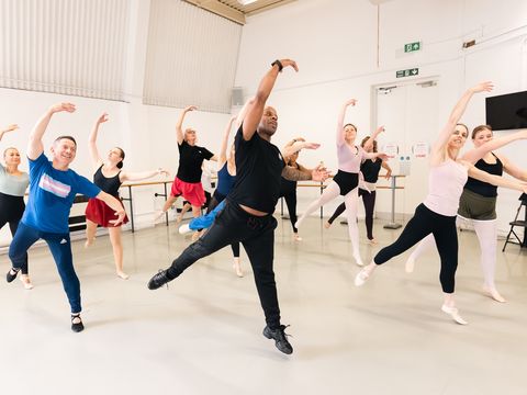 A group of dancers in a studio practice ballet, each striking a graceful pose with arms raised, following a lead instructor in dynamic action.