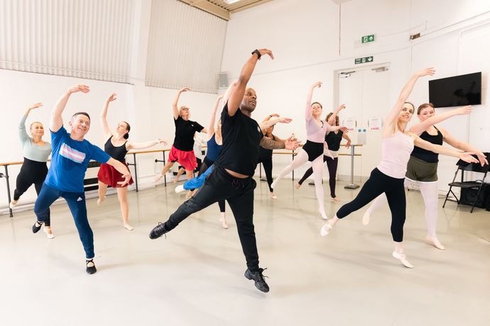 A group of dancers in a studio practice ballet, each striking a graceful pose with arms raised, following a lead instructor in dynamic action.